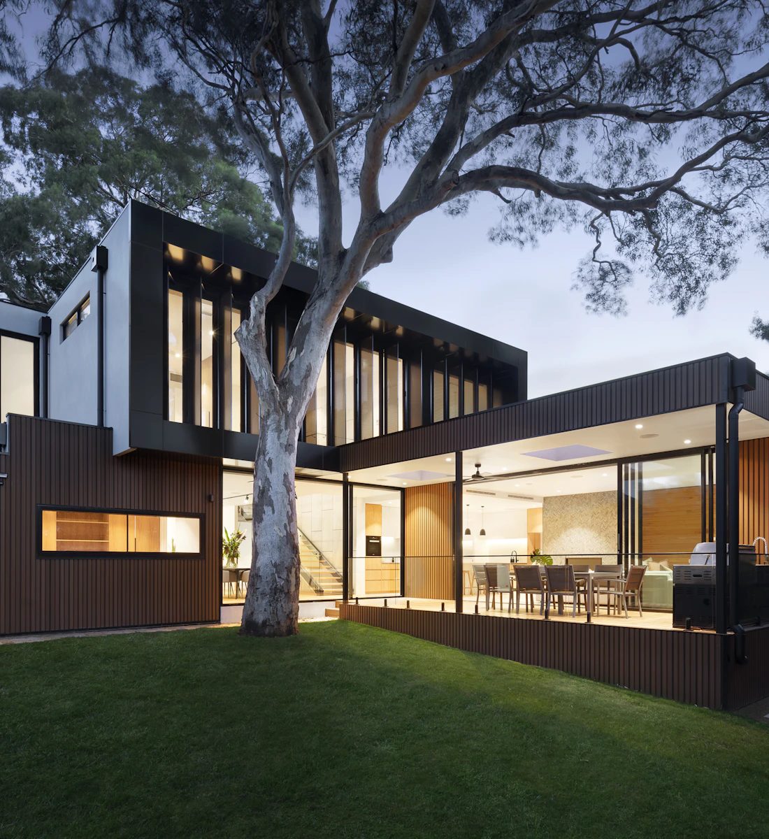 Modern two-story home with lit windows and a large tree on the lawn at dusk